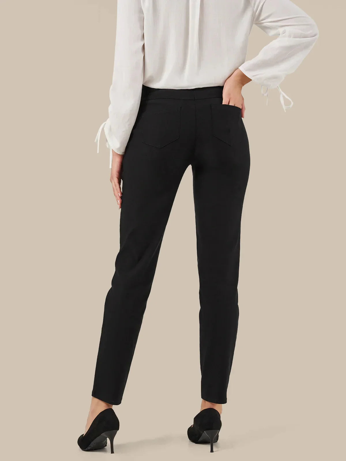 A model from the back wearing slim-fit black trousers, a white long-sleeve blouse with tie details at the cuffs, and black high heels, set against a solid beige background.