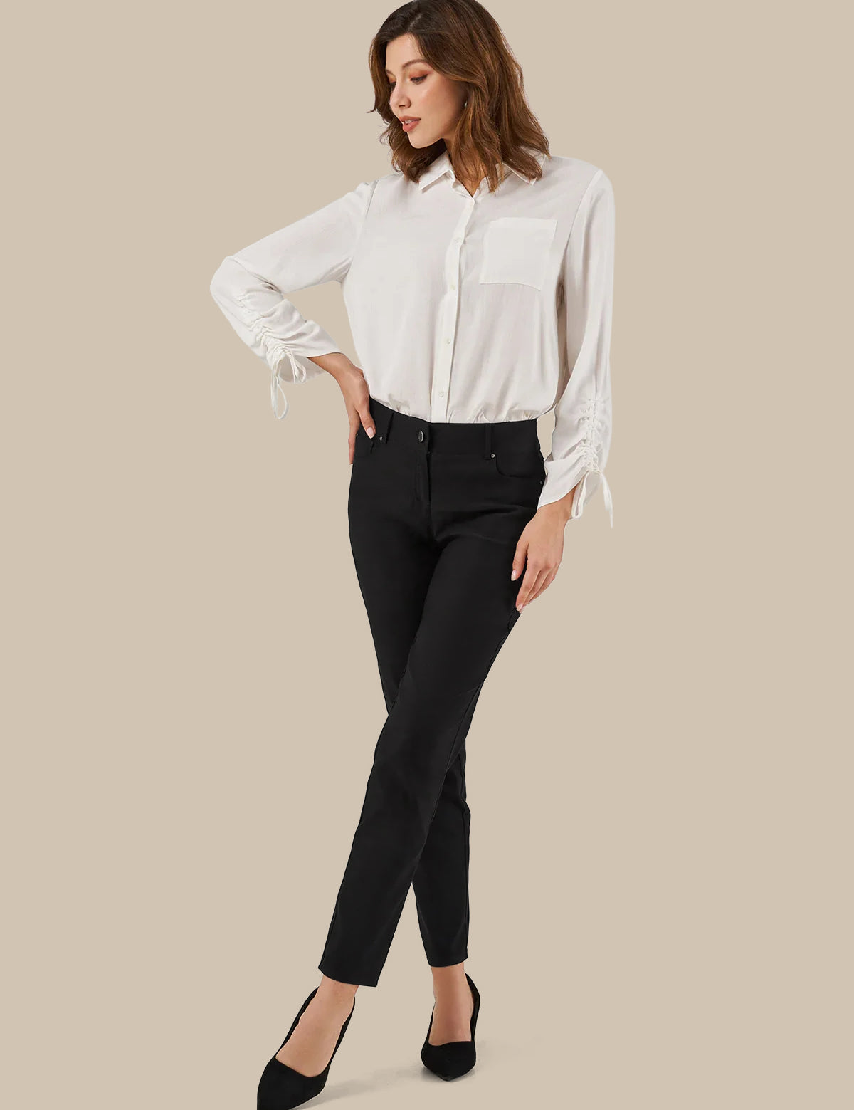 A woman modeling a white long-sleeve button-up blouse with tie details at the cuffs, paired with black ankle-length pants and black pointed-toe heels, against a solid beige background.