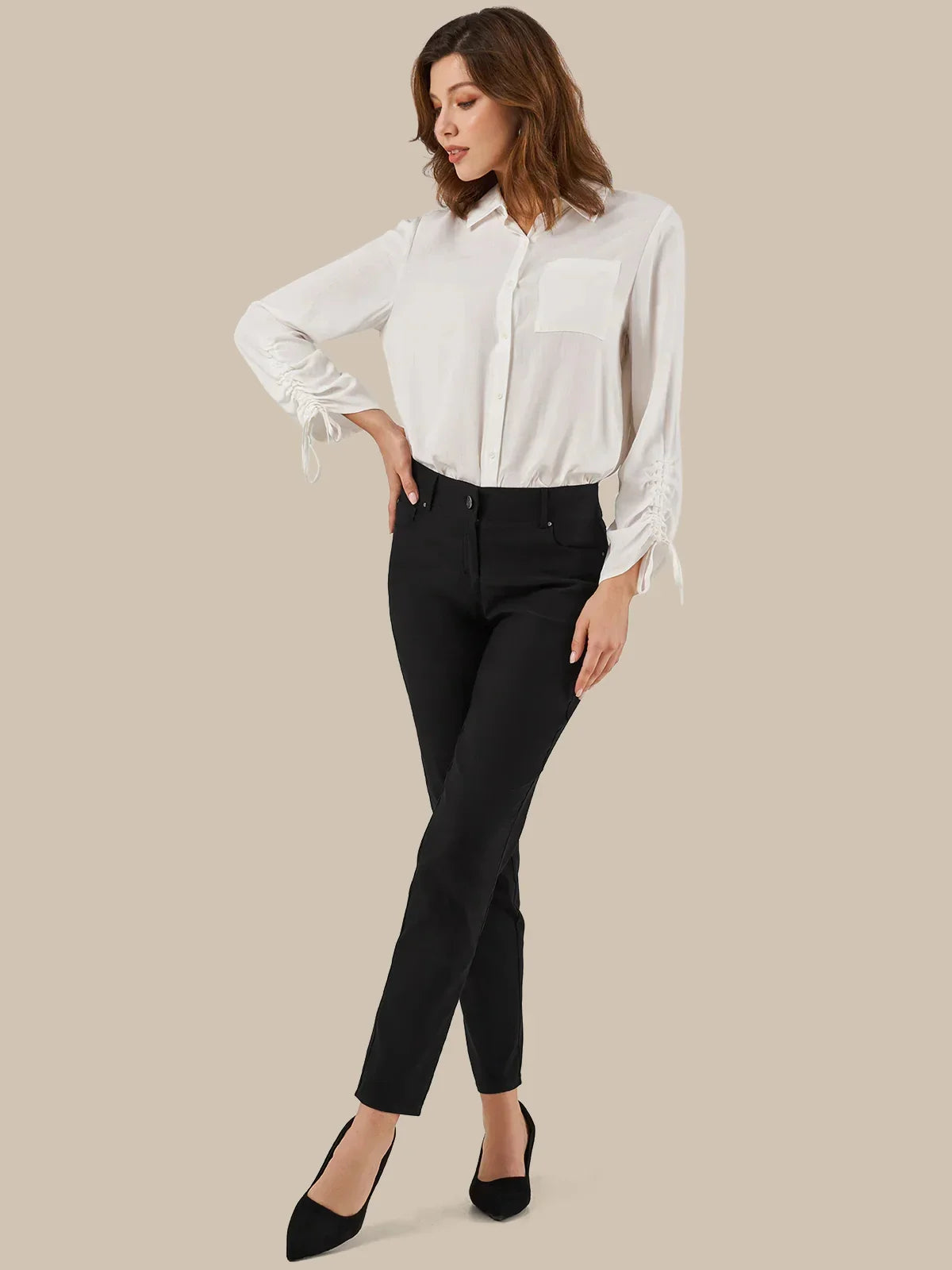 A woman modeling a white long-sleeve button-up blouse with tie details at the cuffs, paired with black ankle-length pants and black pointed-toe heels, against a solid beige background.