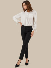 A woman modeling a white long-sleeve button-up blouse with tie details at the cuffs, paired with black ankle-length pants and black pointed-toe heels, against a solid beige background.