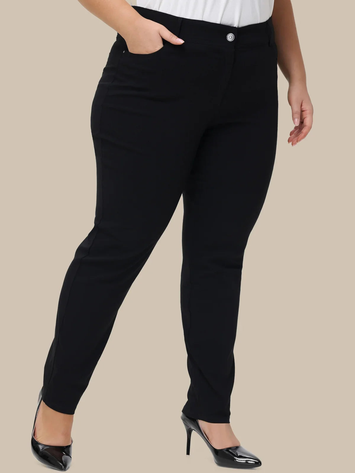 Black slim-fit pants shown on a model from the waist down, paired with a white top and black patent high heels, against a solid beige background.