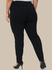 A person shown from the back wearing slim black pants with a white top and black pointed heels, against a plain beige studio background.