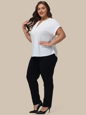 A woman modeling a white short-sleeve V-neck top with black pants and black heels, standing against a solid beige background.