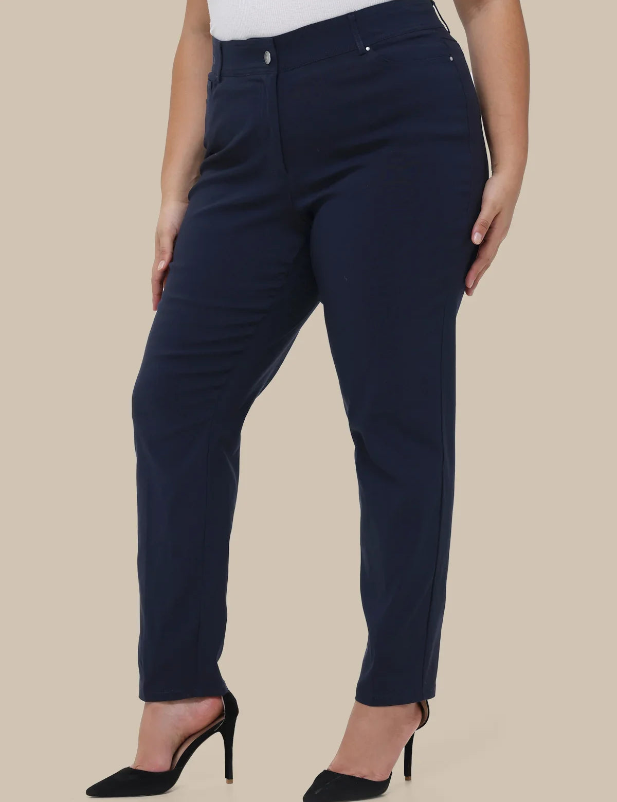 A model wearing navy blue slim-fit trousers with a white top and black high heels, shown from the waist down against a solid beige background.