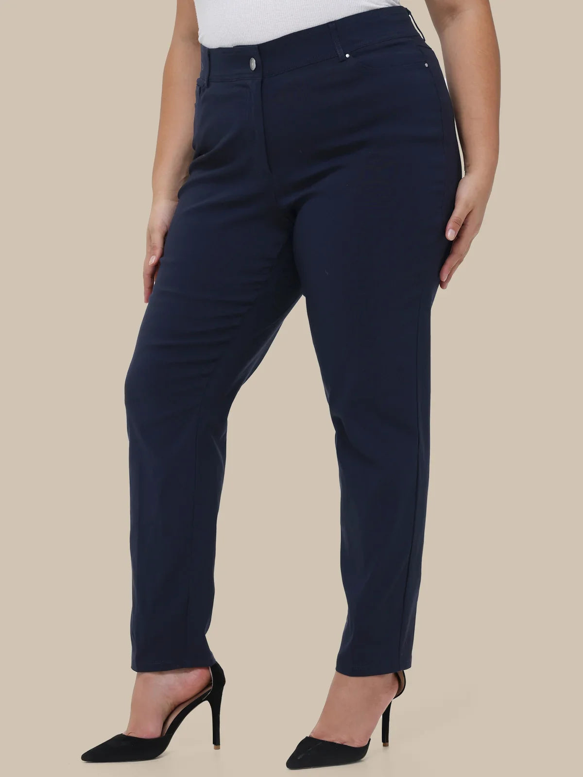 A model wearing navy blue slim-fit trousers with a white top and black high heels, shown from the waist down against a solid beige background.
