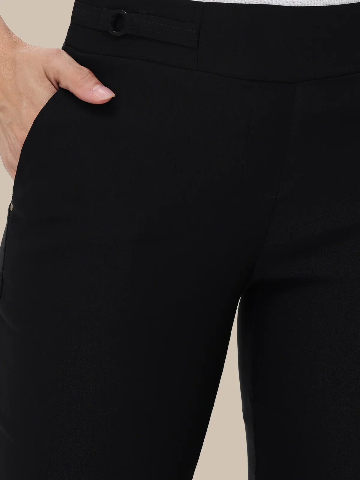 Close-up of a person wearing black tailored trousers, showing the front waistband, side pocket, and hand resting in the pocket against a plain beige background.
