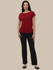 A woman modeling a red short-sleeve top with decorative gold embellishments on the shoulders, paired with black pants and black flats, against a solid beige background.