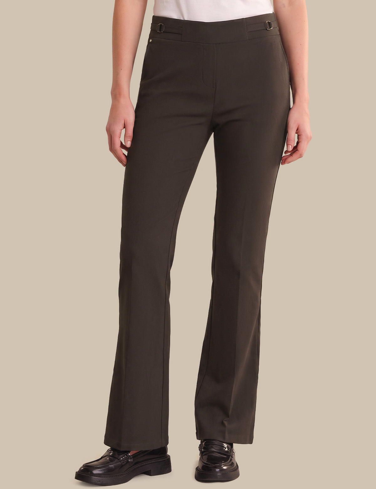 A model wearing dark brown straight-leg trousers, a white top, and black leather loafers, standing against a solid beige background.