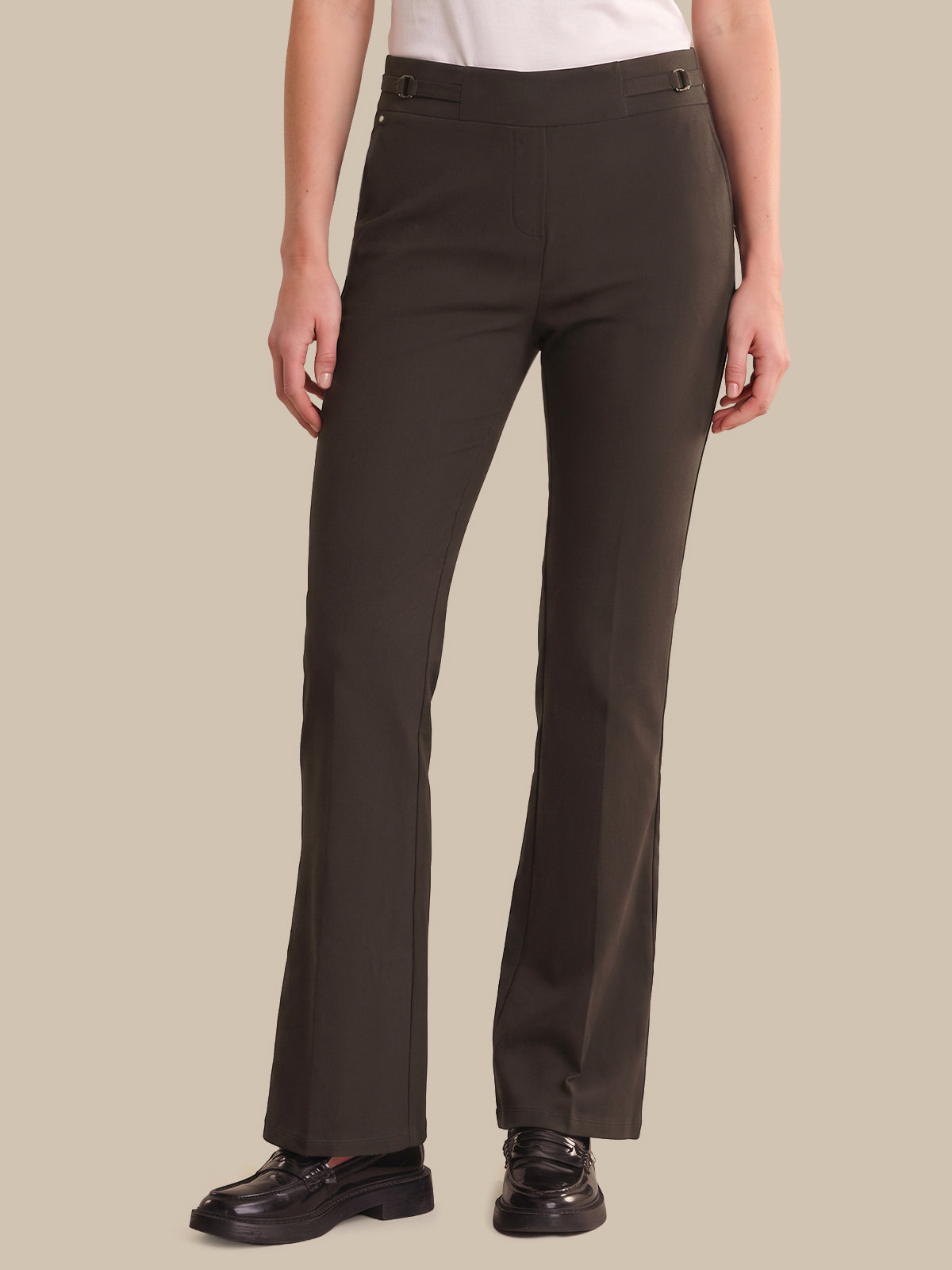 A model wearing dark brown straight-leg trousers, a white top, and black leather loafers, standing against a solid beige background.