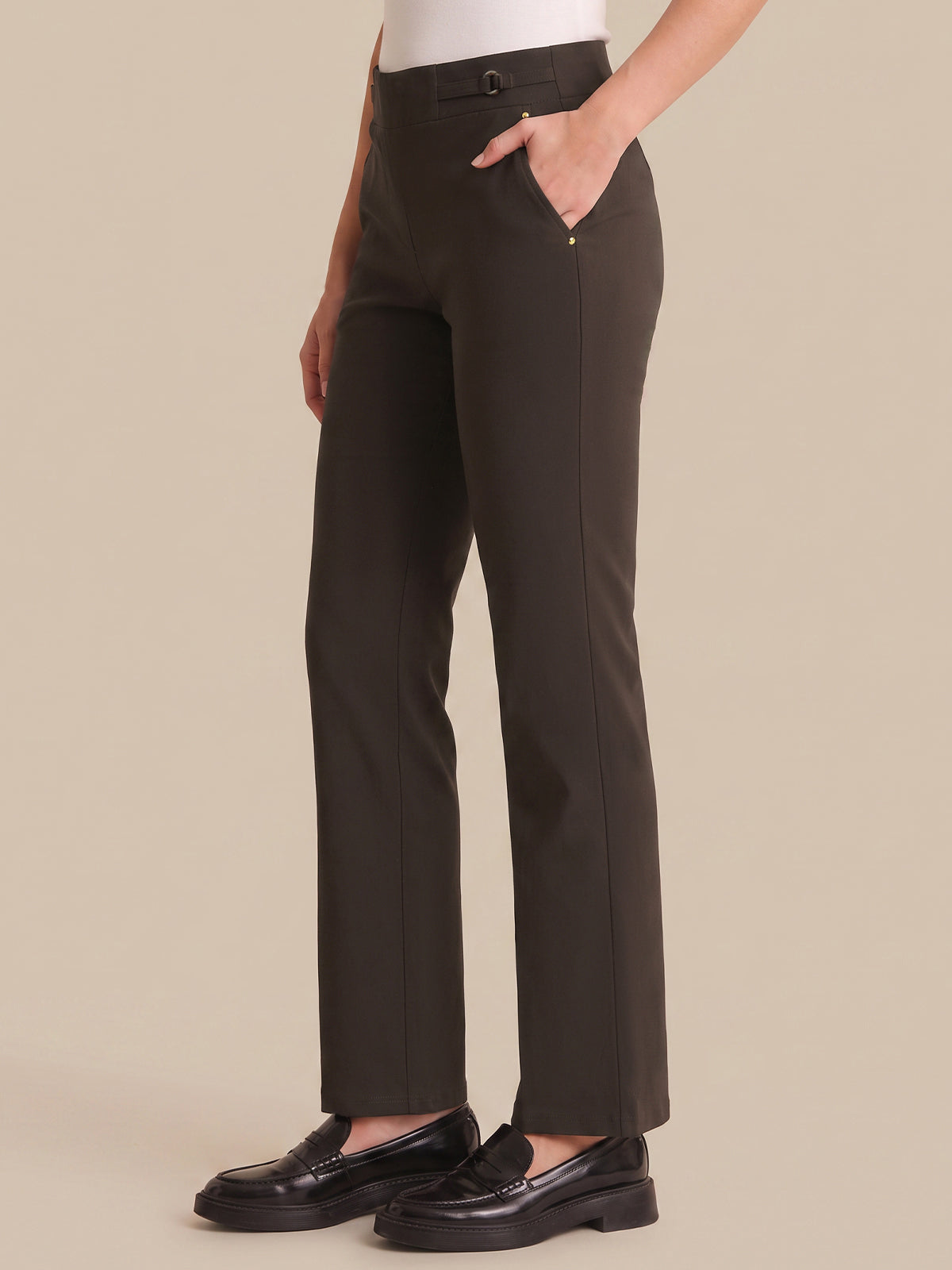 A person wearing dark olive straight-leg trousers with a white top and black loafers, shown from the waist down against a solid beige background.