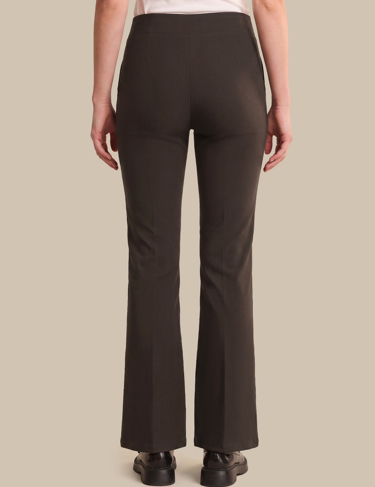 A person shown from the back wearing dark brown flared pants, a light top, and black shoes against a plain beige studio background.