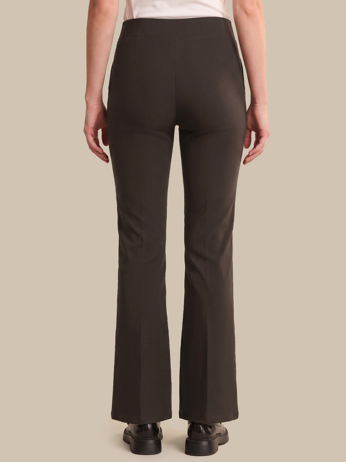 A person shown from the back wearing dark brown flared pants, a light top, and black shoes against a plain beige studio background.