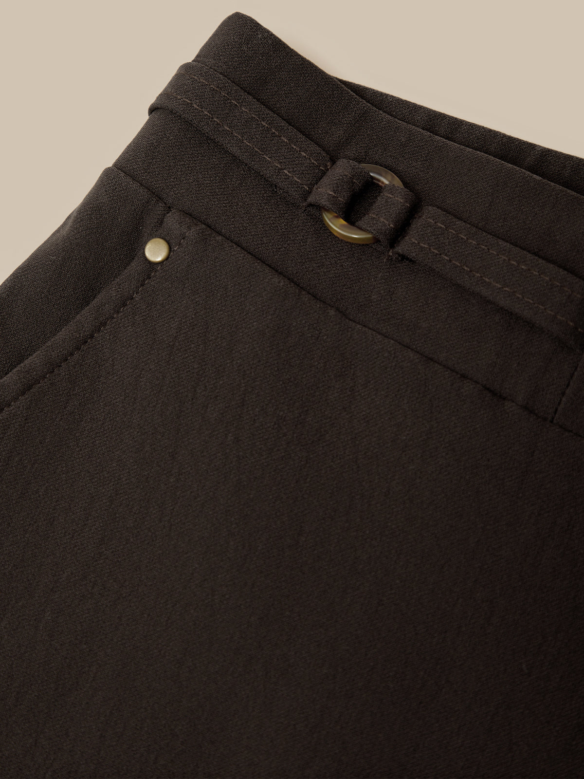 Close-up of a dark brown or very deep espresso-colored fabric garment, showing the waistband with a matching belt and brass-tone metal hardware against a light beige background.