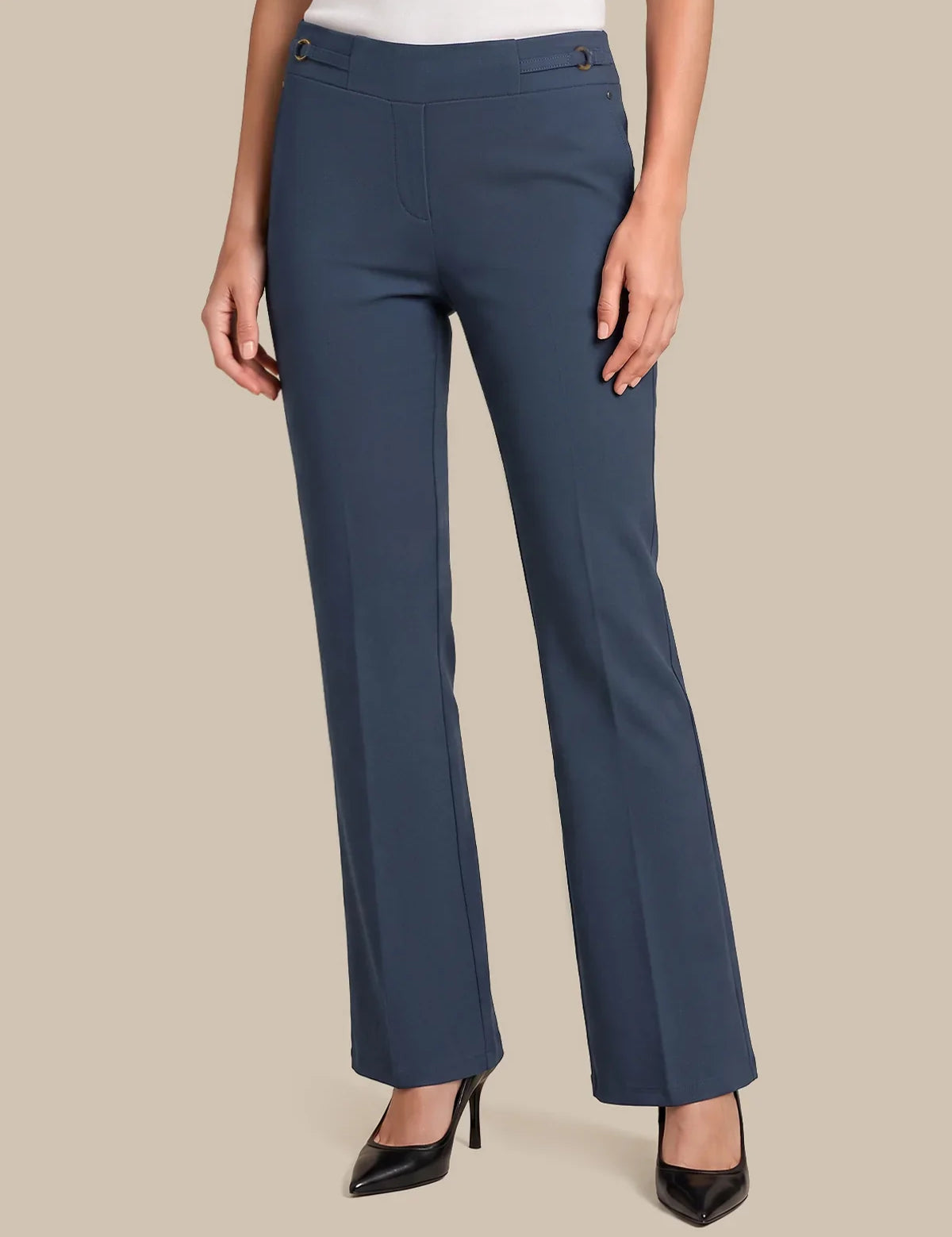 A model wearing a pair of slate blue straight-leg trousers with a white top and black pointed-toe heels, shown from the waist down against a solid beige background.