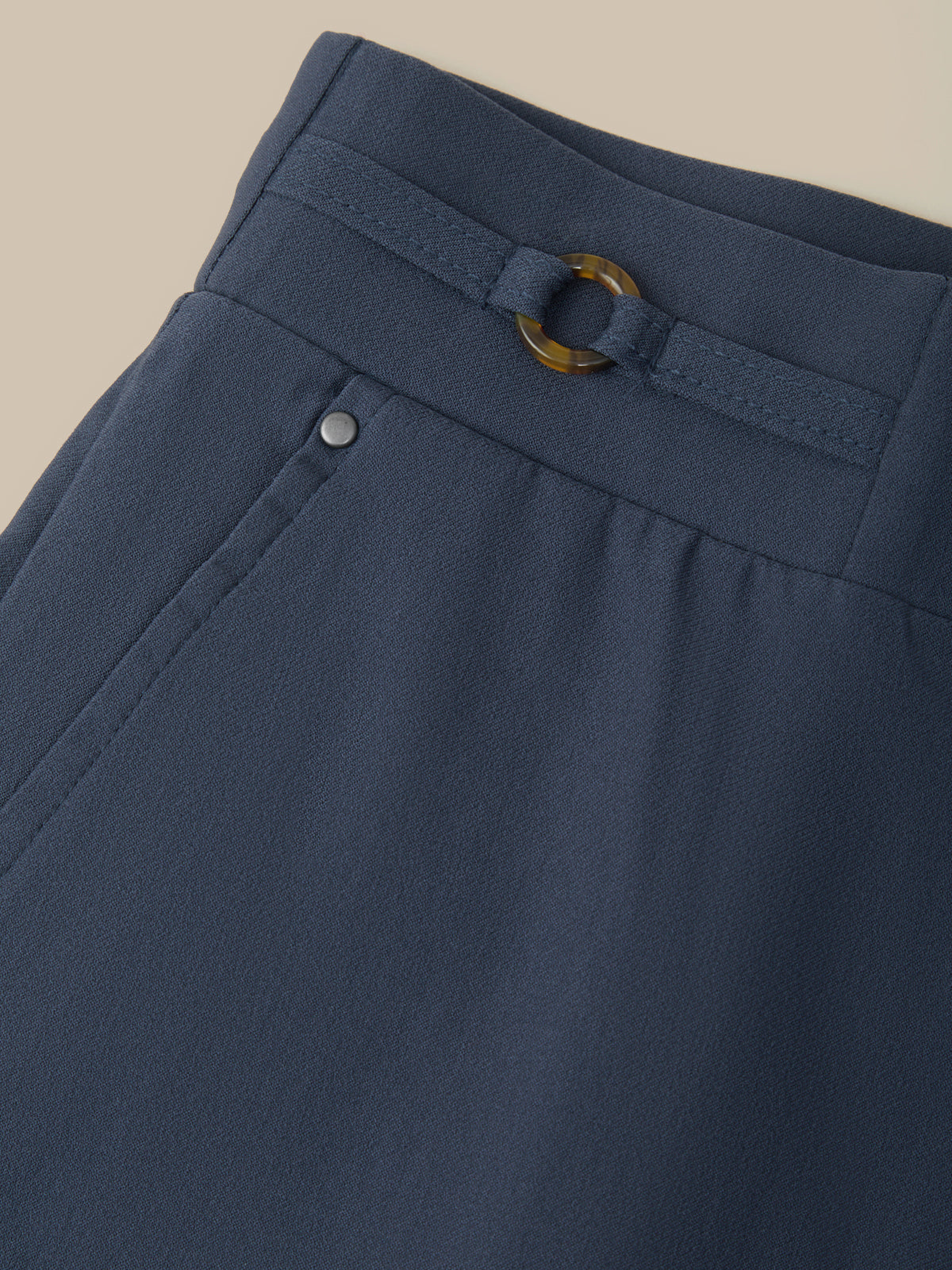 Close-up of navy blue trousers showing the waistband with a matching belt and a gold-tone ring buckle, plus a small silver button detail.