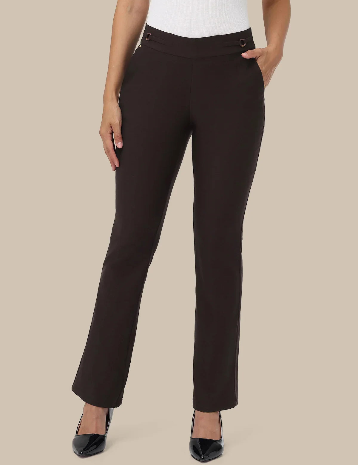 A model wearing dark brown straight-leg trousers with a double button waistband, styled with a white top and black pointed-toe heels against a solid beige background.