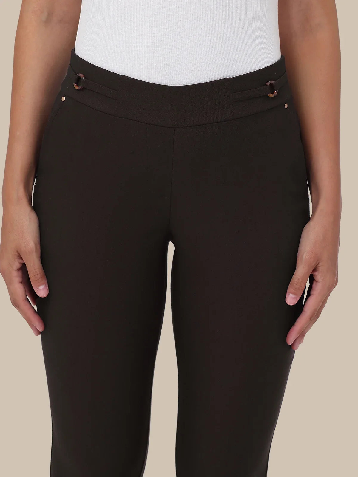 A close-up of a model wearing dark brown high-waisted riding pants with decorative brown grommet details at the waistband, paired with a white ribbed top against a solid beige background.
