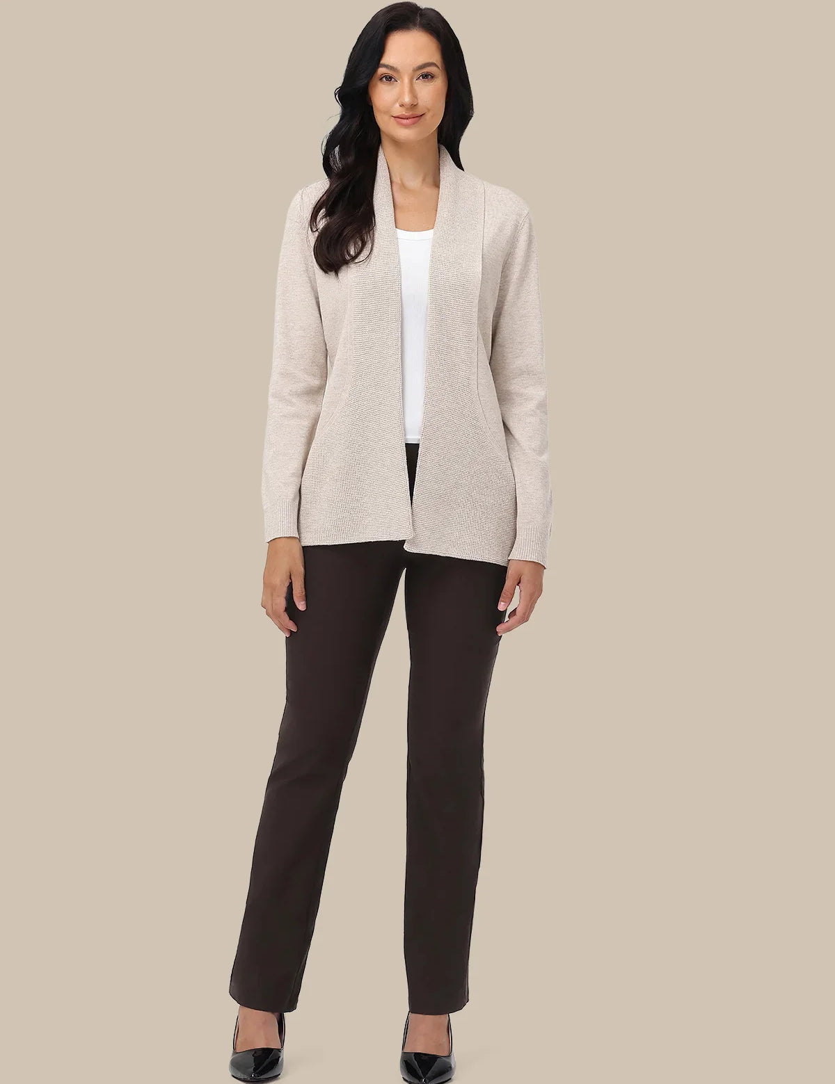 A woman modeling a light beige open-front cardigan over a white top and dark brown pants, standing against a solid beige background.