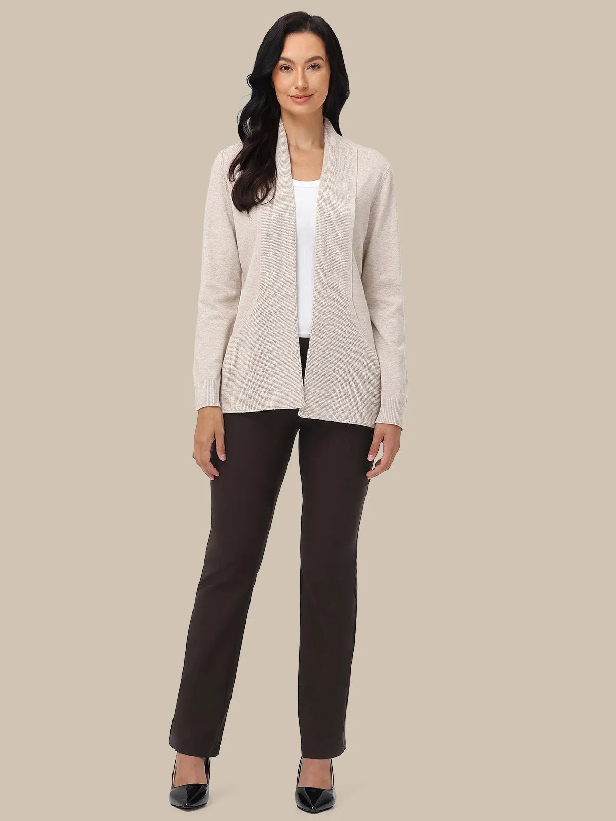 A woman modeling a light beige open-front cardigan over a white top and dark brown pants, standing against a solid beige background.