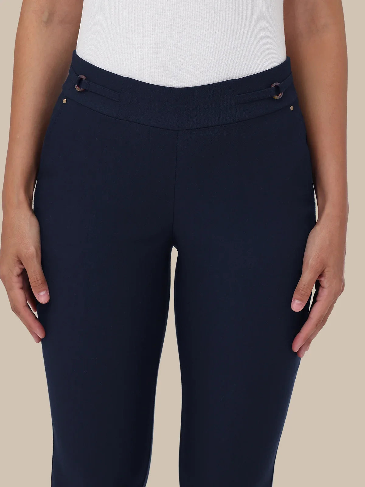 A close-up of a model wearing navy blue riding breeches with decorative brown and gold buttons on the waistband, paired with a white ribbed top against a solid beige background.