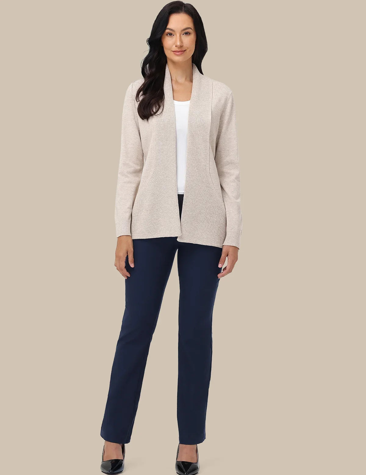 A woman modeling a light beige open-front knit cardigan over a white top and navy blue pants, standing against a solid beige studio background.