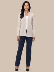 A woman modeling a light beige open-front knit cardigan over a white top and navy blue pants, standing against a solid beige studio background.