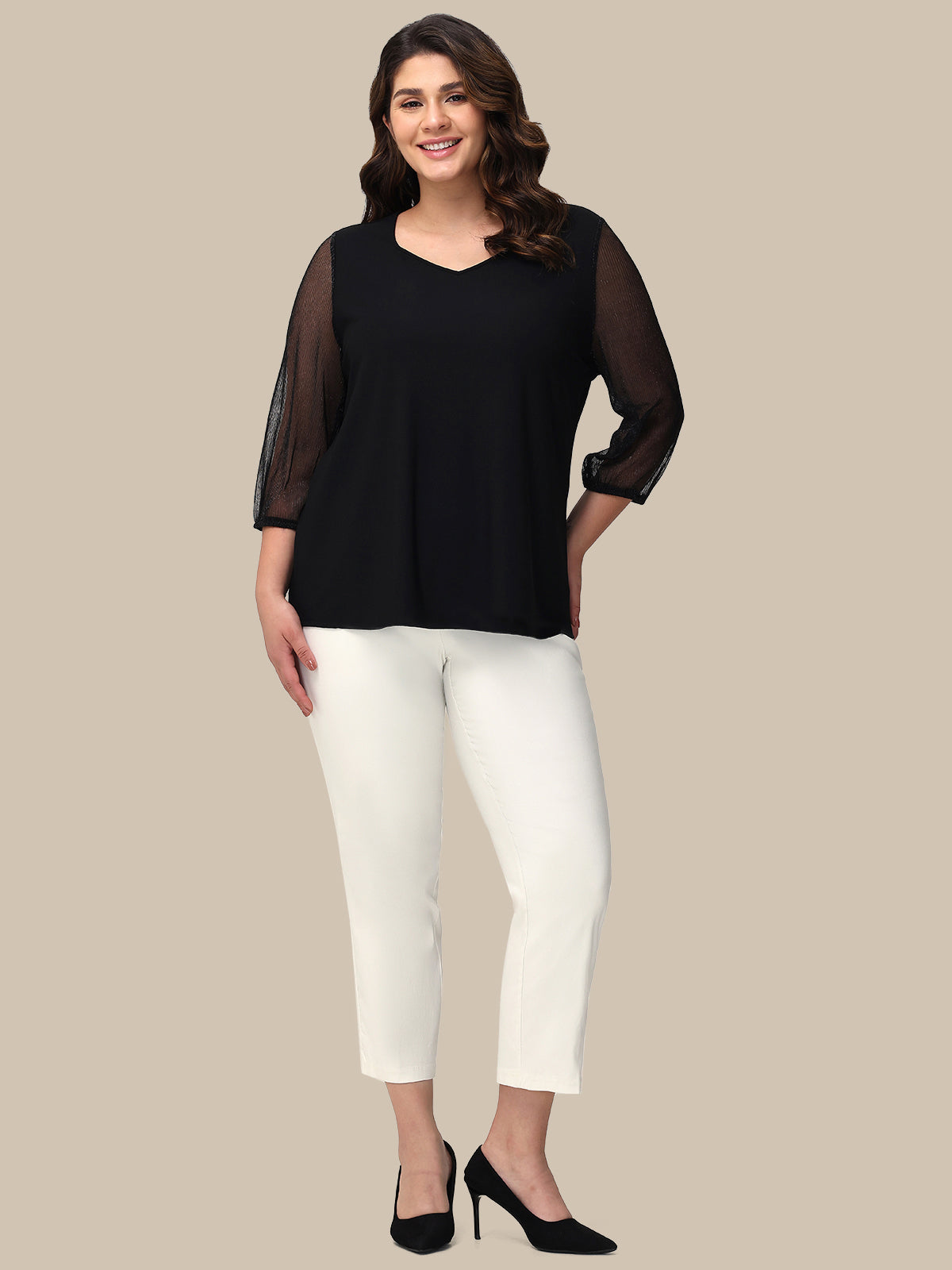 Woman wearing a black top with sheer sleeves and white pants on a beige background