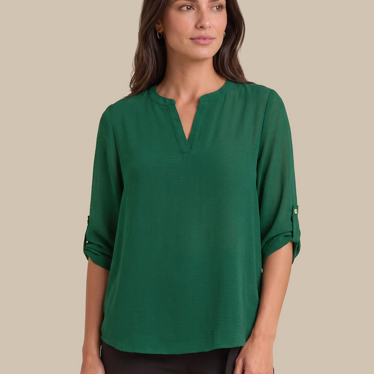 A woman modeling a green long-sleeve blouse with a split V-neckline and rolled sleeves, paired with black pants, against a solid beige background.