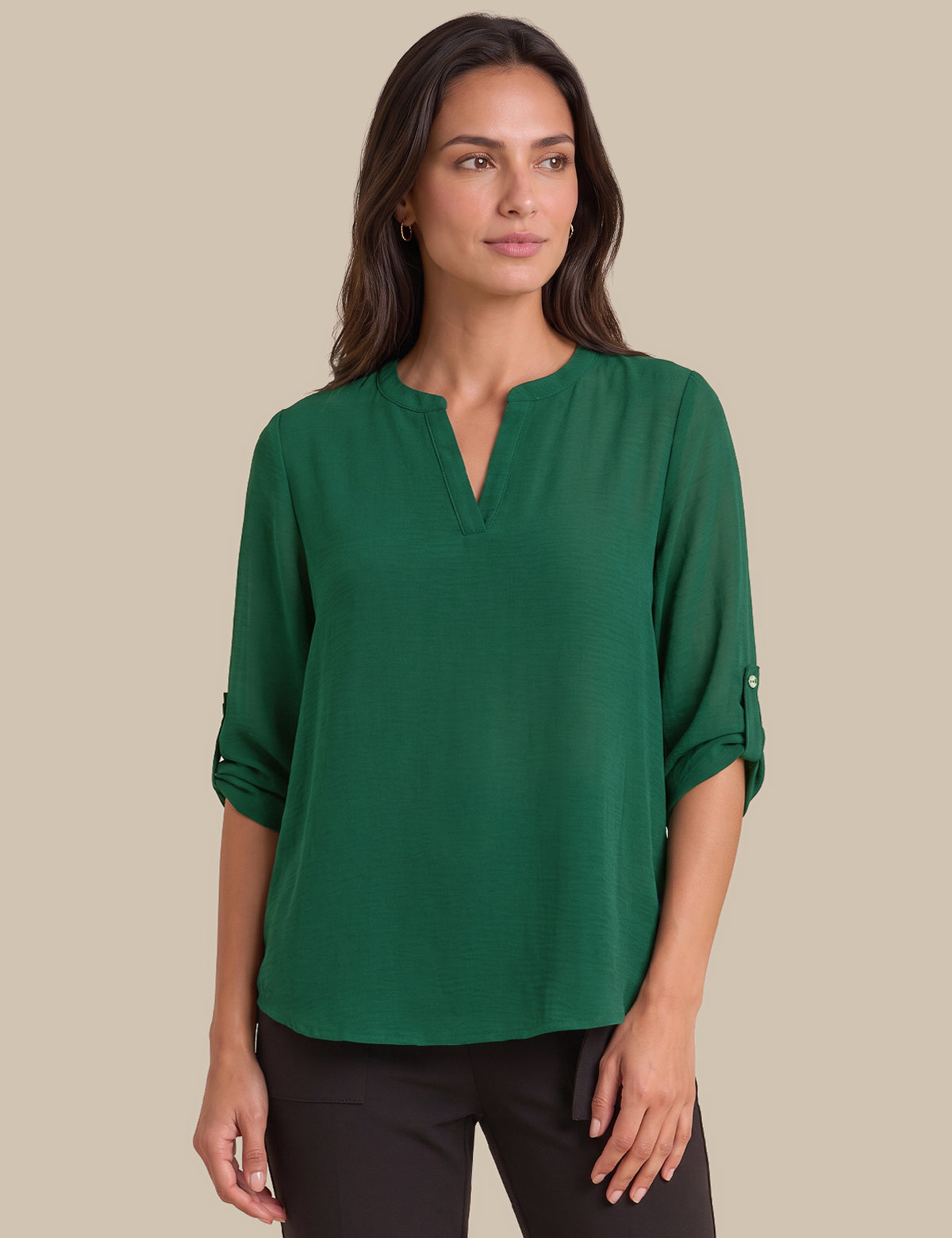 A woman modeling a green long-sleeve blouse with a split V-neckline and rolled sleeves, paired with black pants, against a solid beige background.