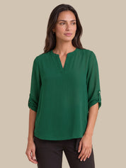A woman modeling a green long-sleeve blouse with a split V-neckline and rolled sleeves, paired with black pants, against a solid beige background.