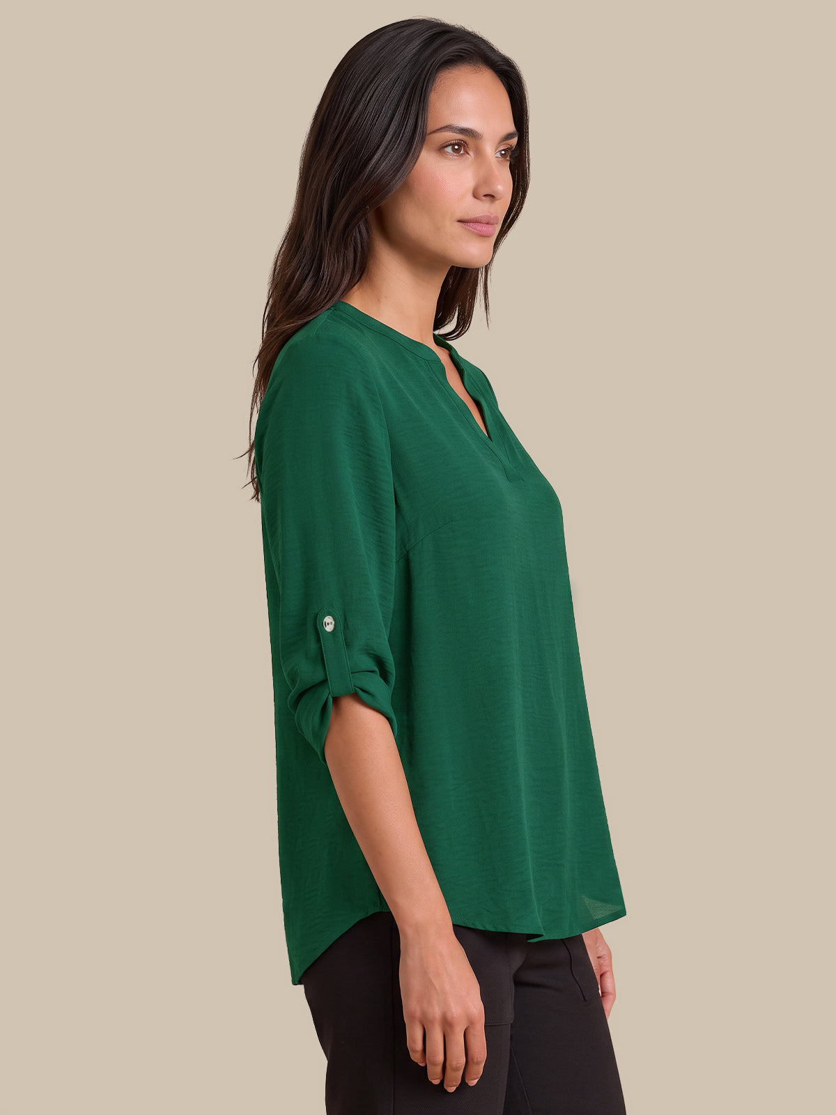 A woman wearing a green V-neck top with rolled three-quarter sleeves and black pants, photographed against a solid beige studio background.