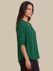 A woman wearing a green V-neck top with rolled three-quarter sleeves and black pants, photographed against a solid beige studio background.