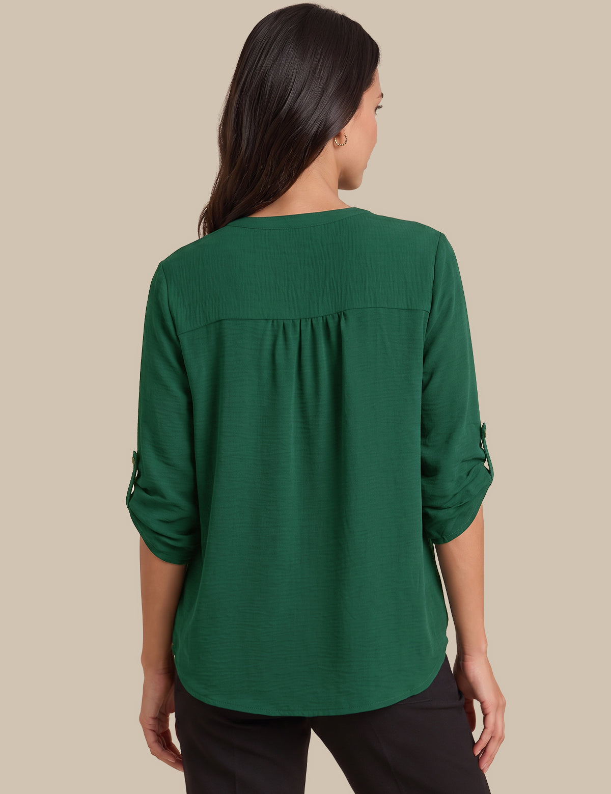 A woman viewed from the back wearing a dark green long-sleeve blouse with rolled cuffs and black pants, set against a solid beige background.