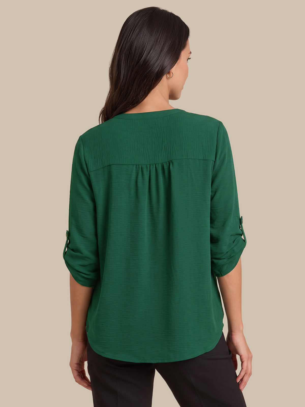 A woman viewed from the back wearing a dark green long-sleeve blouse with rolled cuffs and black pants, set against a solid beige background.