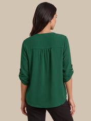 A woman viewed from the back wearing a dark green long-sleeve blouse with rolled cuffs and black pants, set against a solid beige background.