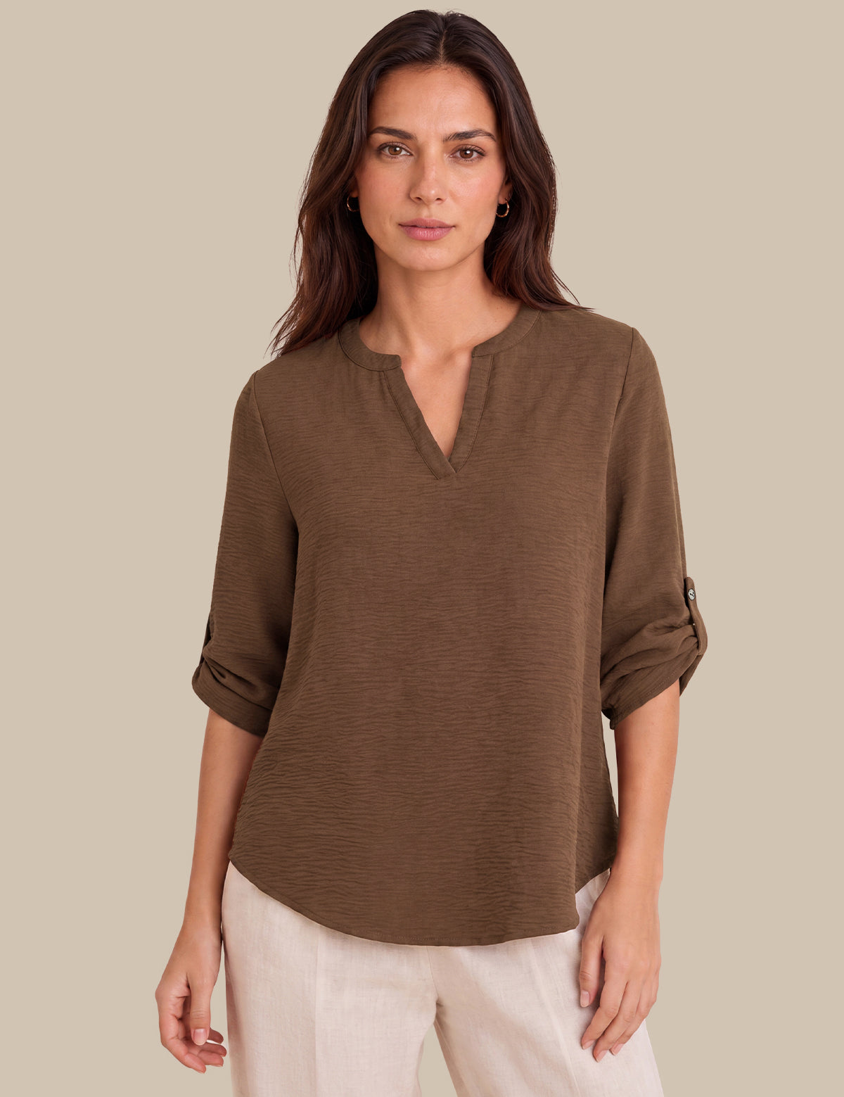 A woman modeling a brown, long-sleeve, henley-style top with rolled-up sleeves, paired with light beige pants, against a solid beige background.