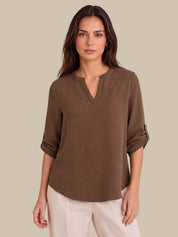 A woman modeling a brown, long-sleeve, henley-style top with rolled-up sleeves, paired with light beige pants, against a solid beige background.