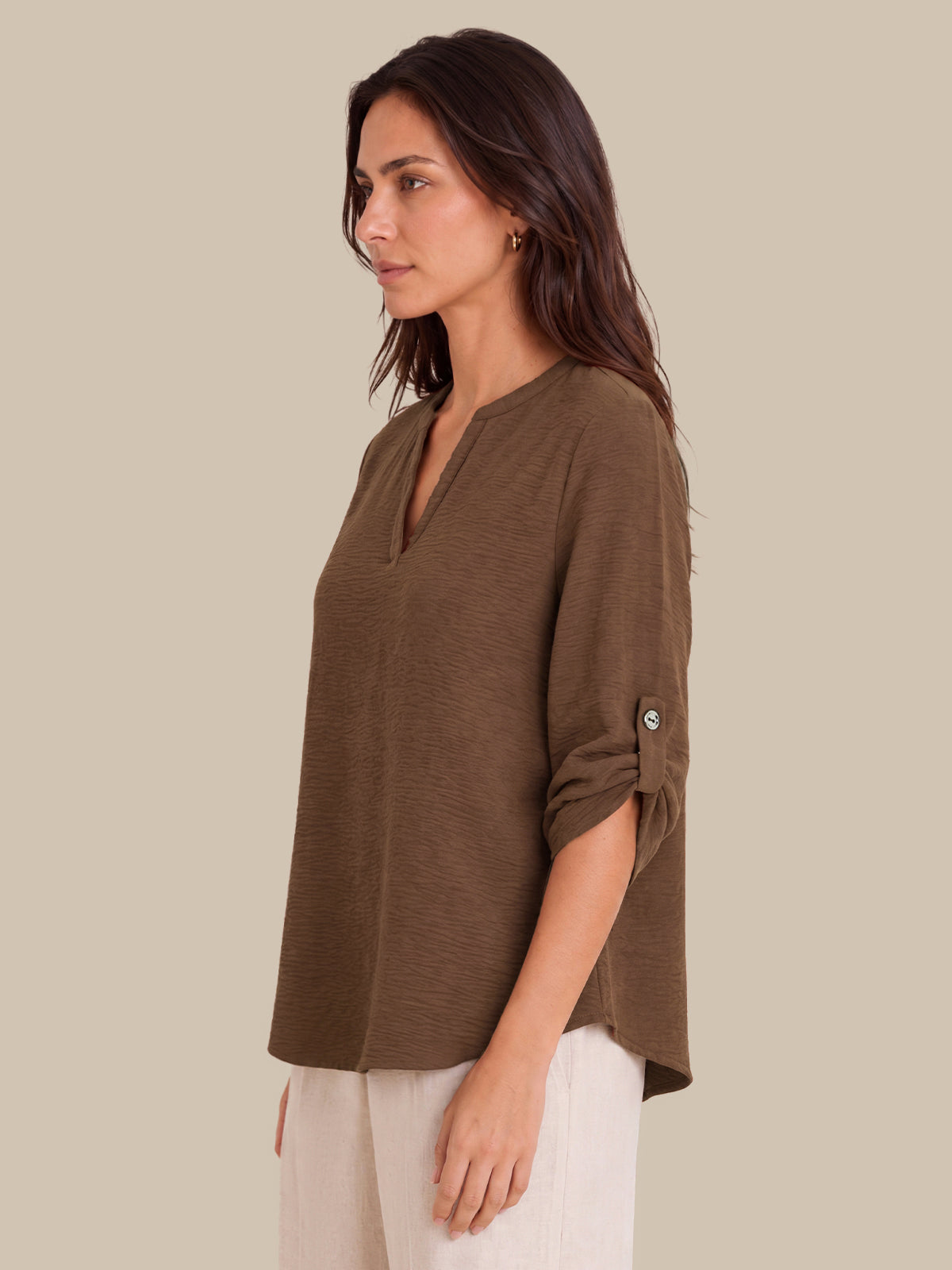 A woman shown in side profile wearing a loose brown V-neck top with rolled sleeves and light-colored pants, photographed against a plain beige studio background.