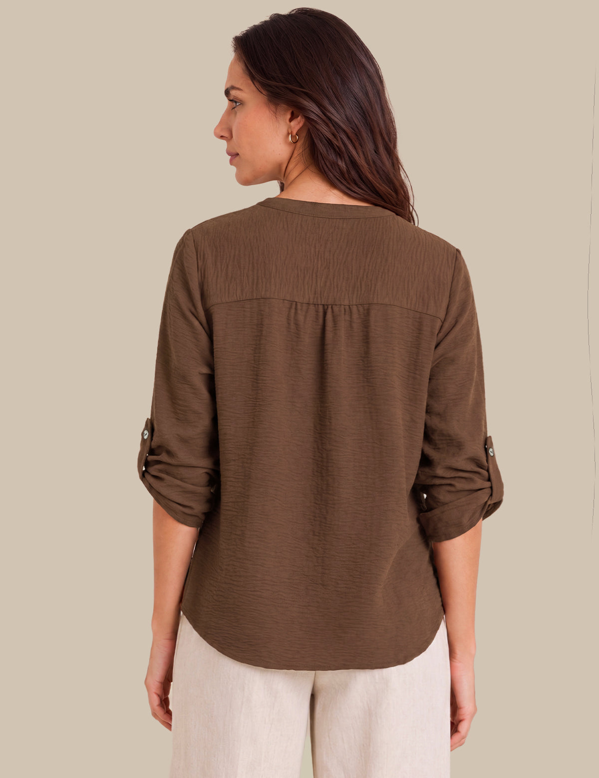 A woman shown from the back wearing a brown long-sleeve blouse with rolled sleeves and light beige pants, set against a solid beige background.