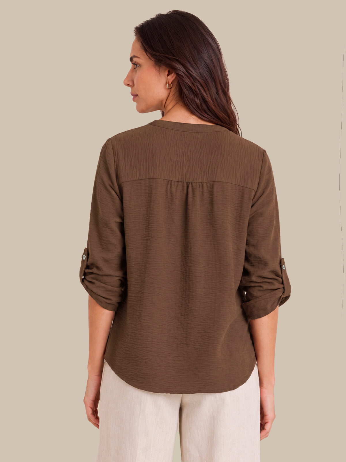 A woman shown from the back wearing a brown long-sleeve blouse with rolled sleeves and light beige pants, set against a solid beige background.