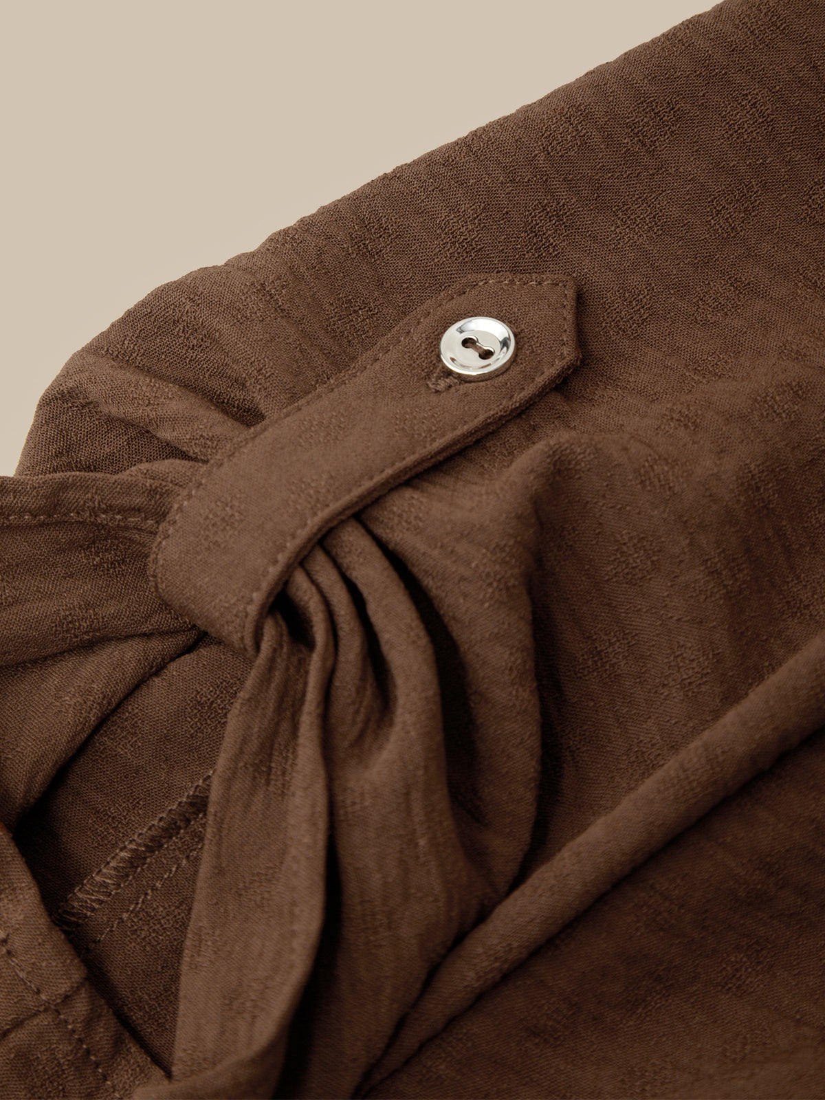 Close-up of a brown textured garment with a tie detail and a small silver button, shown against a plain beige background.