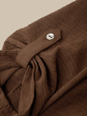 Close-up of a brown textured garment with a tie detail and a small silver button, shown against a plain beige background.