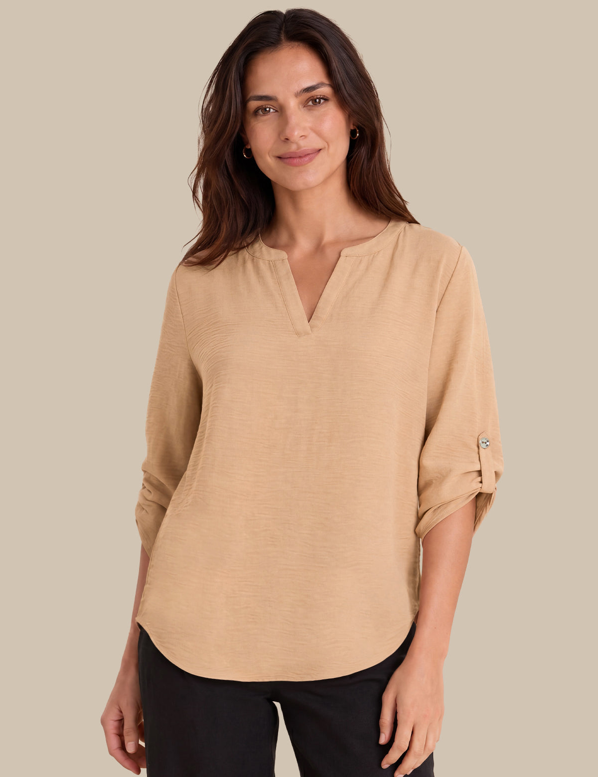 A woman modeling a beige long-sleeve top with a split V-neckline and rolled sleeves, paired with black pants, against a solid beige background.