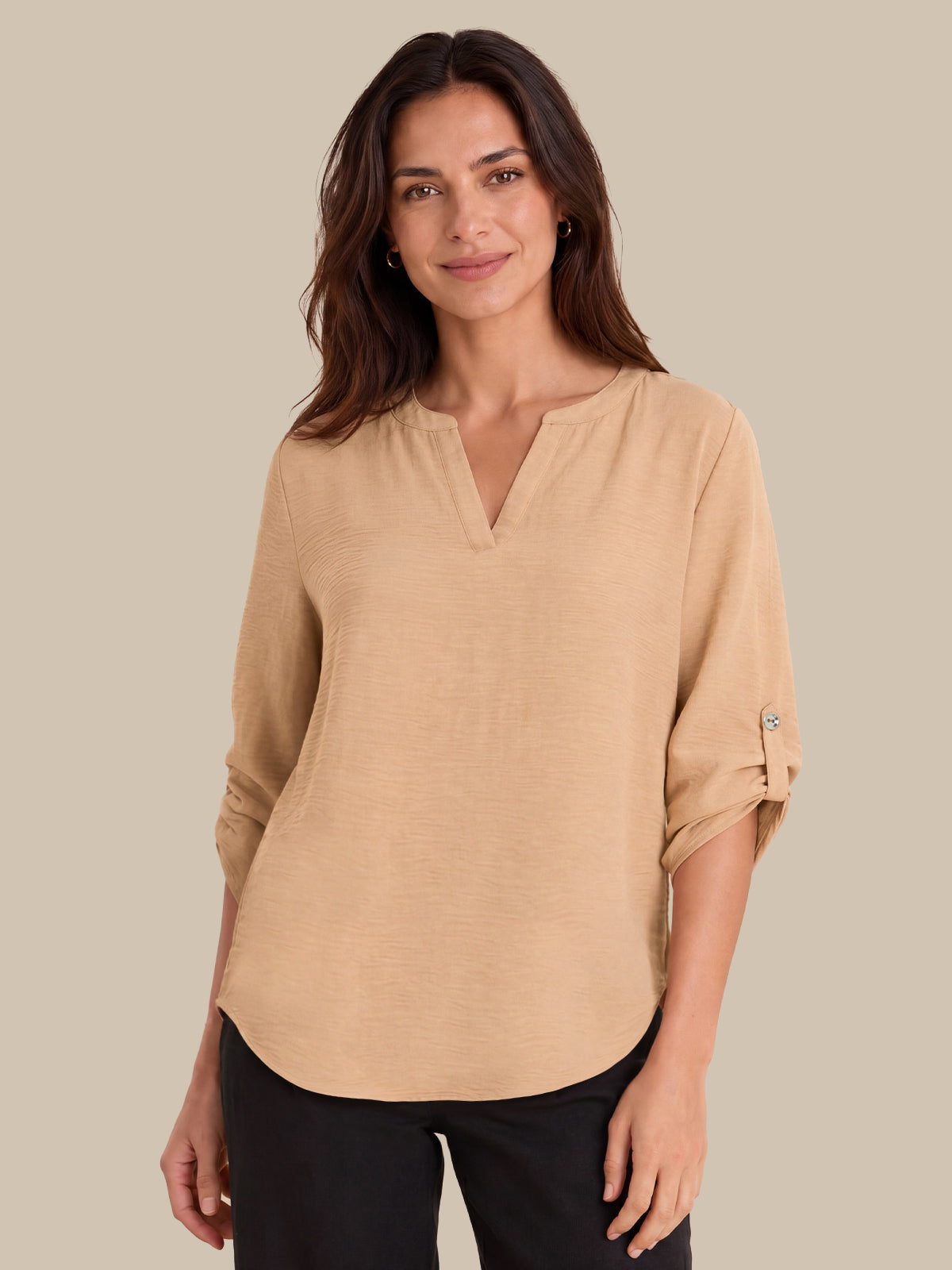 A woman modeling a beige long-sleeve top with a split V-neckline and rolled sleeves, paired with black pants, against a solid beige background.