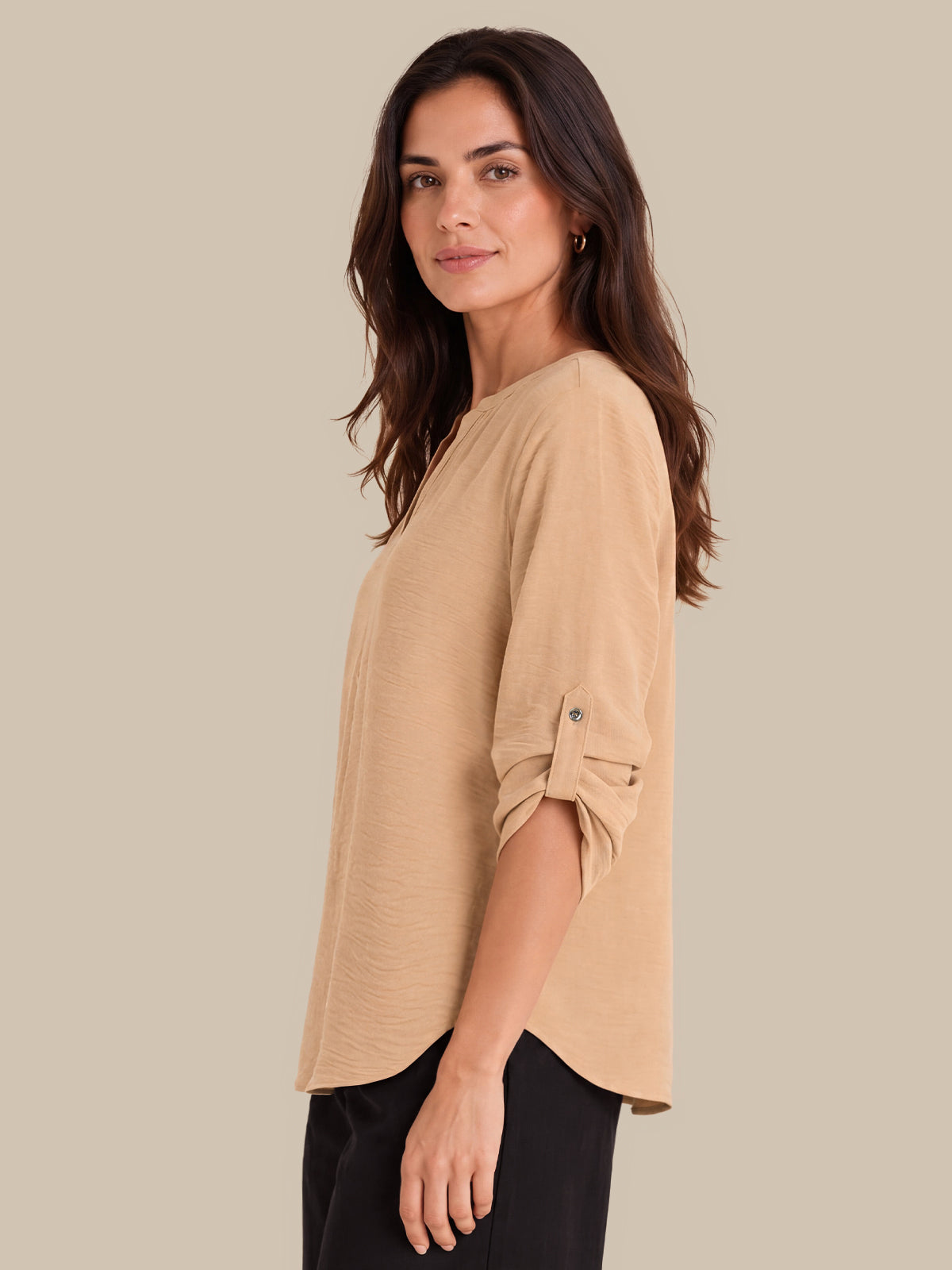 A woman wearing a beige, relaxed-fit top with rolled sleeves and black pants, photographed against a solid beige background.