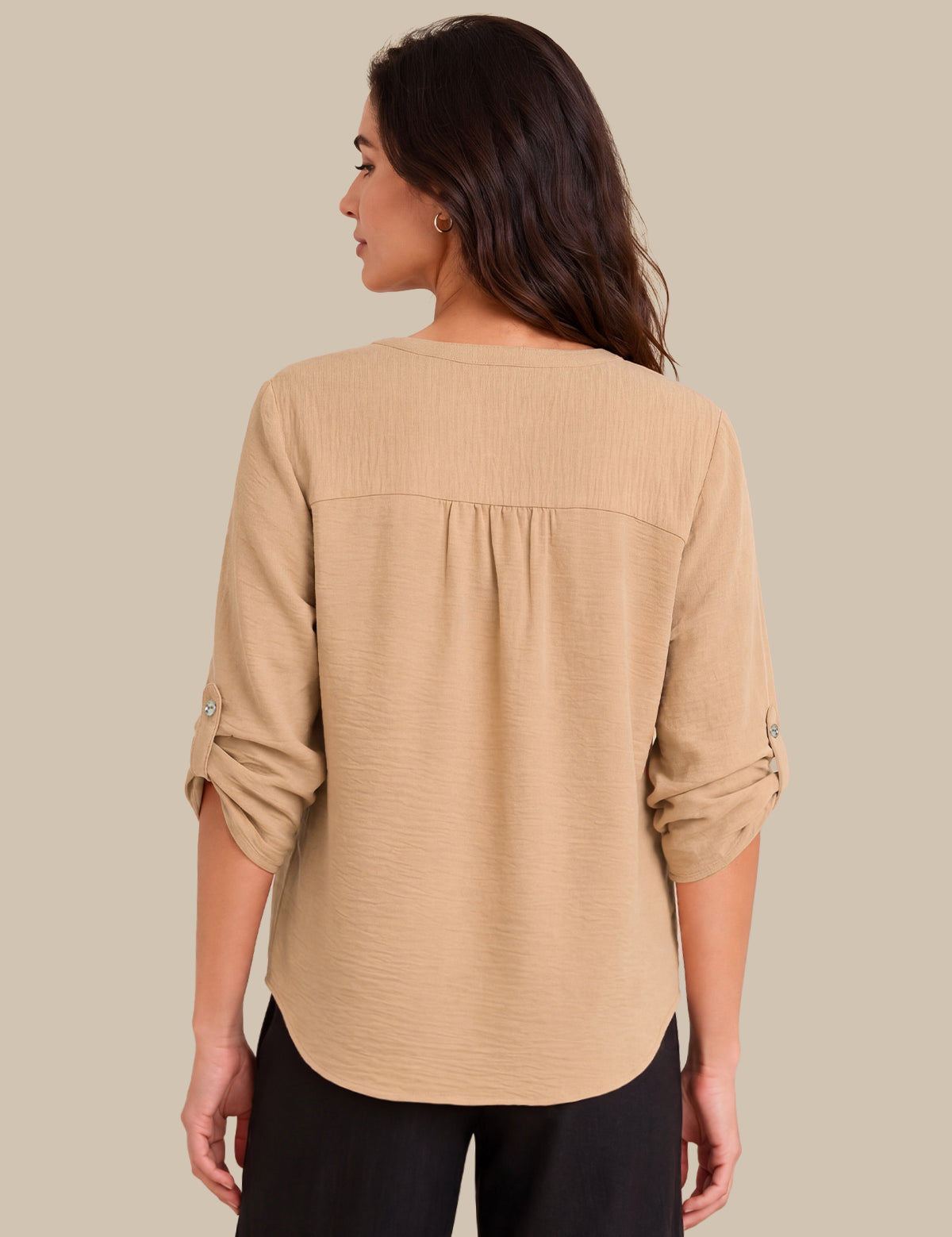 A woman shown from the back wearing a beige textured long-sleeve blouse with gathered detailing at the shoulders and rolled sleeves, paired with black pants, against a solid beige background.