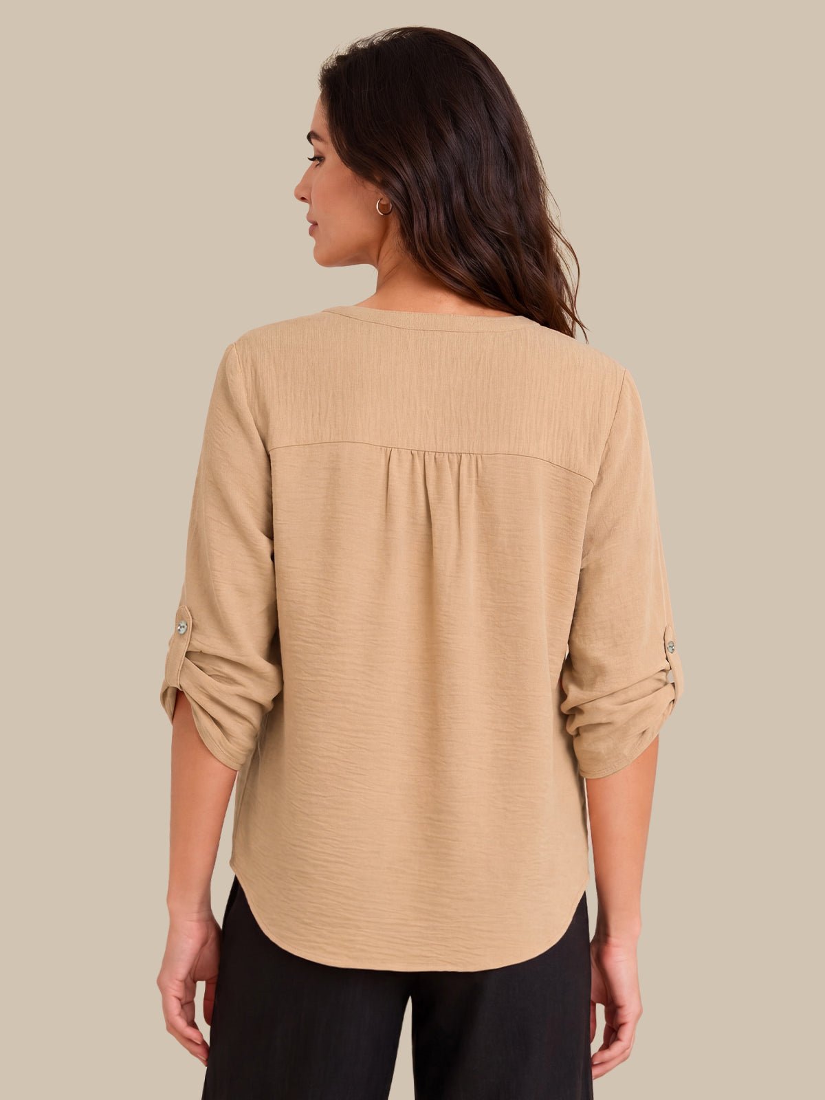 A woman shown from the back wearing a beige textured long-sleeve blouse with gathered detailing at the shoulders and rolled sleeves, paired with black pants, against a solid beige background.