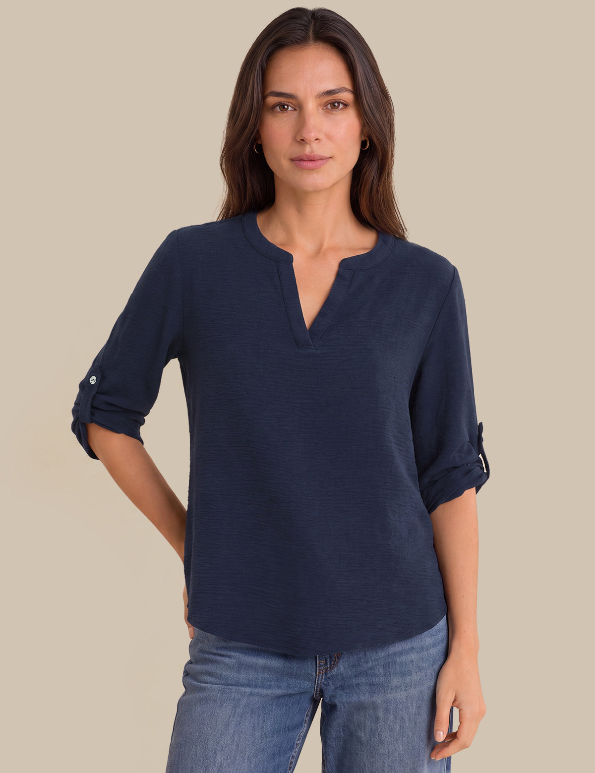 A woman modeling a navy blue long-sleeve henley top with rolled-up sleeves and light blue jeans, set against a solid beige background.
