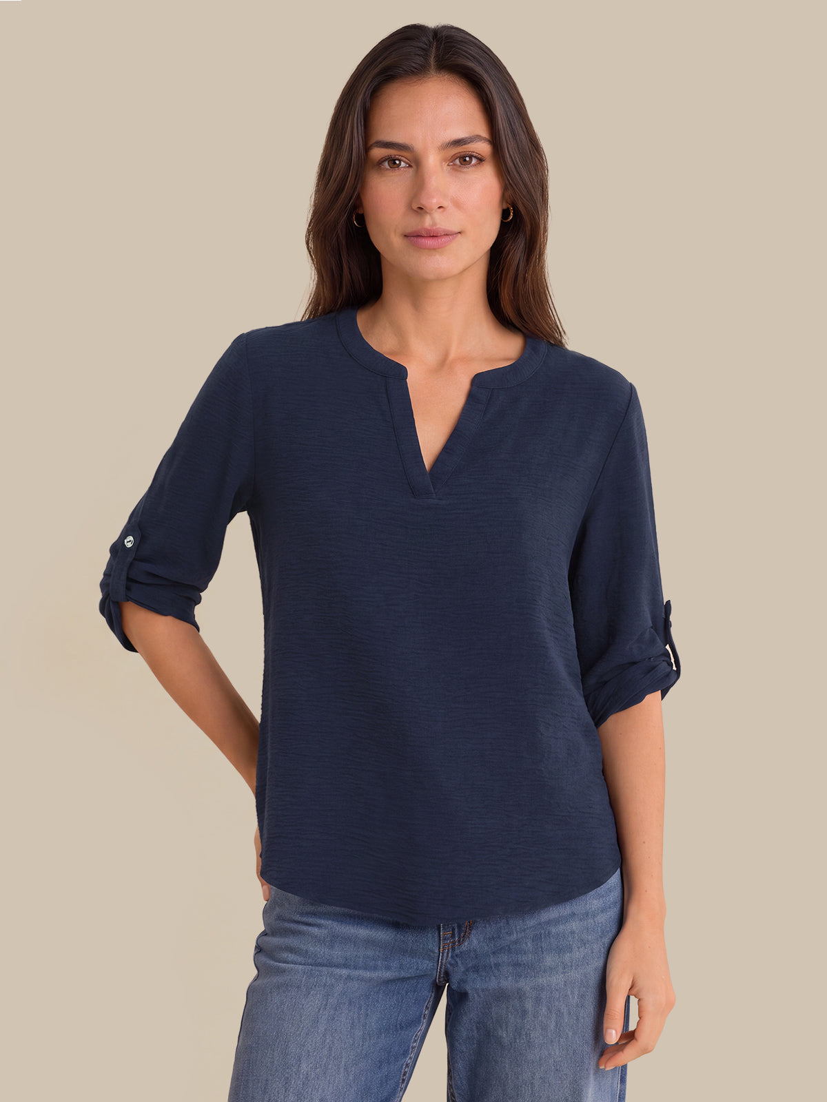 A woman modeling a navy blue long-sleeve henley top with rolled-up sleeves and light blue jeans, set against a solid beige background.