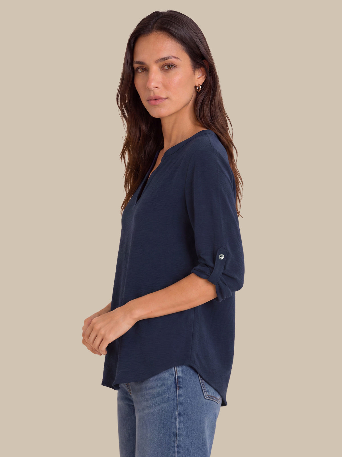 A woman wearing a navy blue V-neck top with rolled-up sleeves and blue jeans, standing against a solid beige background.